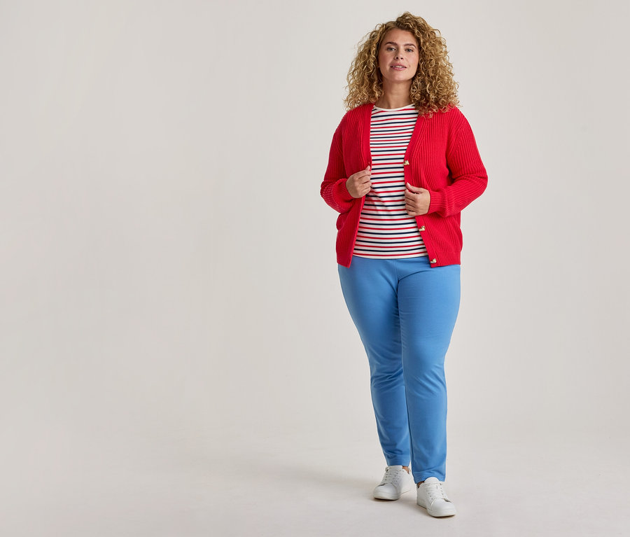 Une femme aux cheveux bouclés se tient debout, vêtue d'un cardigan rouge déboutonné sur un t-shirt rayé, d'un pantalon bleu clair et de baskets blanches.