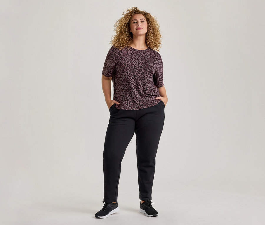 Une femme aux cheveux bouclés se tient de face, vêtue d'un haut à manches courtes imprimé léopard violet foncé, d'un pantalon noir et de baskets noires. Ses mains sont dans ses poches.