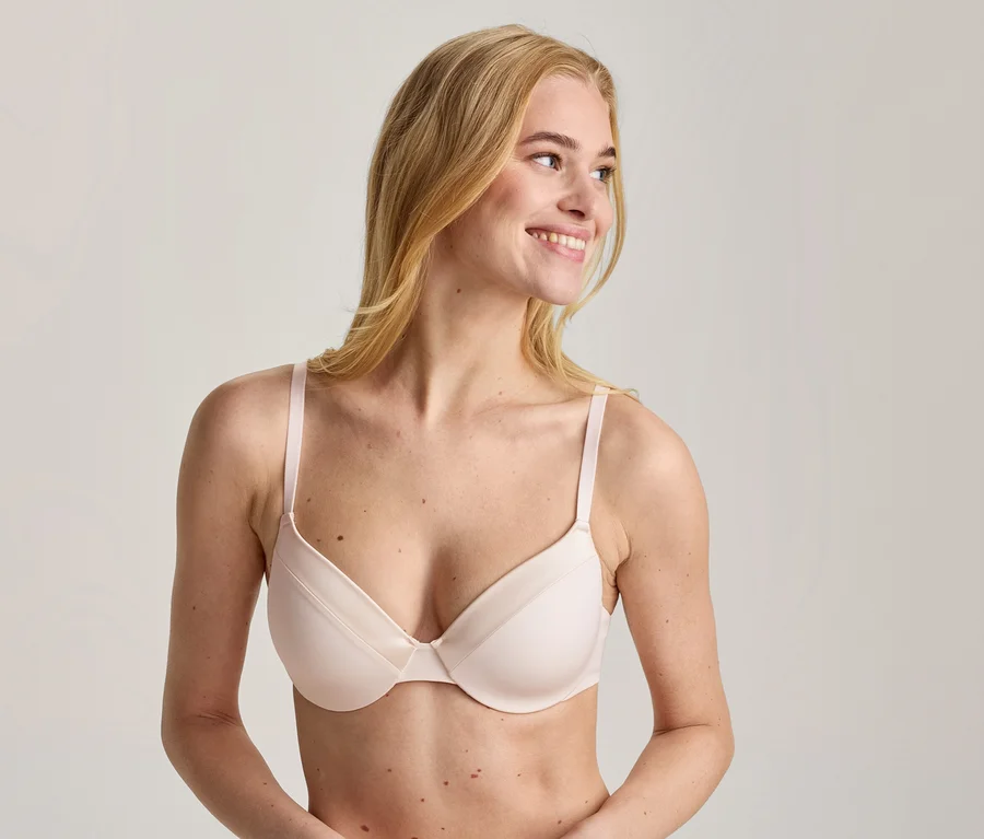 Femme blonde portant un soutien-gorge rose clair, souriant et regardant vers la droite.