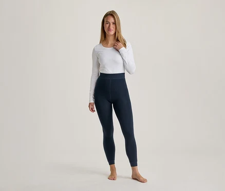 Une femme aux longs cheveux châtains clairs se tient pieds nus, vêtue d'un haut blanc à manches longues et d'un legging bleu foncé, regardant devant elle.