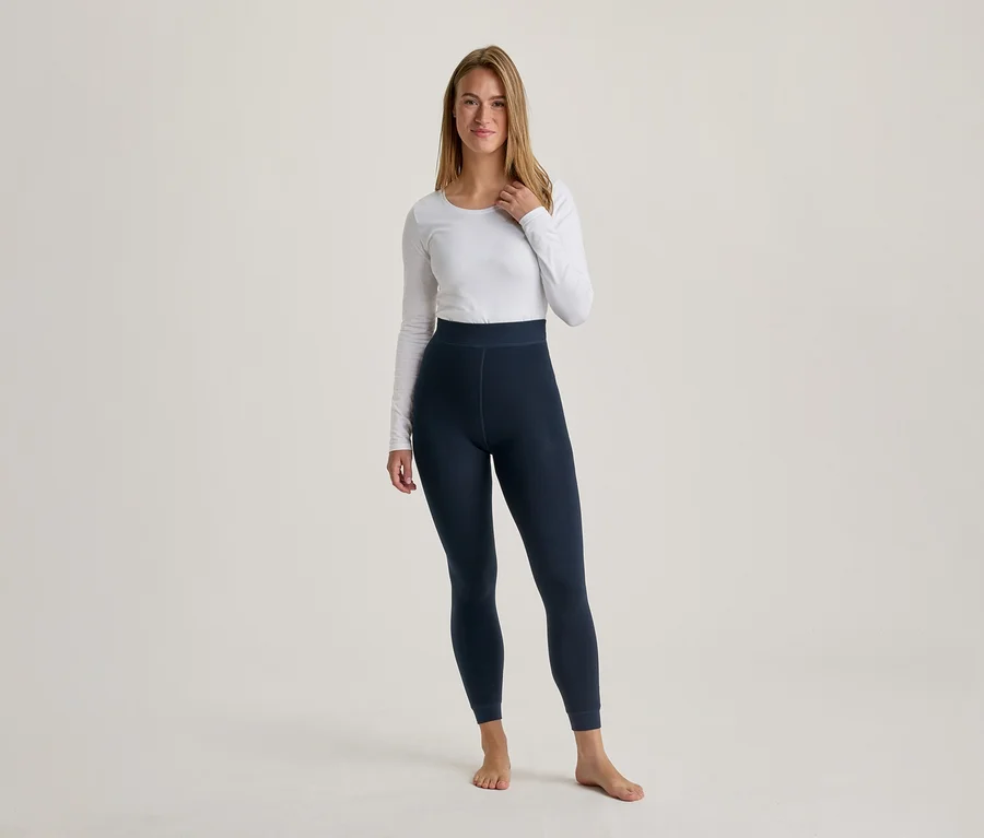 Une femme aux longs cheveux châtains clairs se tient pieds nus, vêtue d'un haut blanc à manches longues et d'un legging bleu foncé, regardant devant elle.