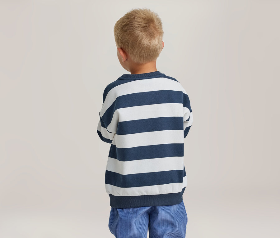 Jeune garçon aux cheveux blonds courts, debout de dos, portant un haut à manches longues rayé bleu marine et blanc et un pantalon bleu.
