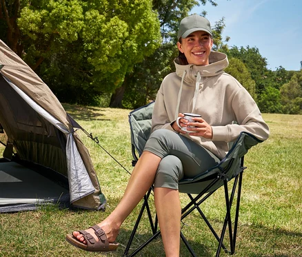 Une femme assise dans une chaise de camping Tambu »ASANA« tient une tasse et porte un legging 3/4 et un sweat à capuche.