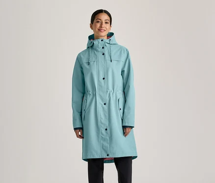 Femme portant un long imperméable bleu clair avec une capuche, des boutons noirs et un cordon de serrage à la taille, sur un pantalon noir.