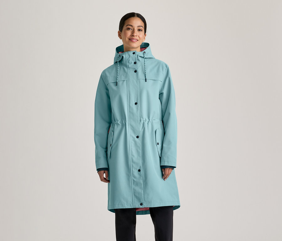 Femme portant un long imperméable bleu clair avec une capuche, des boutons noirs et un cordon de serrage à la taille, sur un pantalon noir.