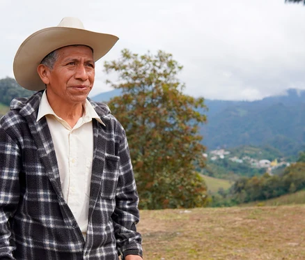 Un homme âgé portant un chapeau de cowboy clair et une veste à carreaux noire et blanche se tient dehors, regardant vers la droite. Derrière lui se trouvent des montagnes vertes, une vallée avec un village et un ciel couvert.