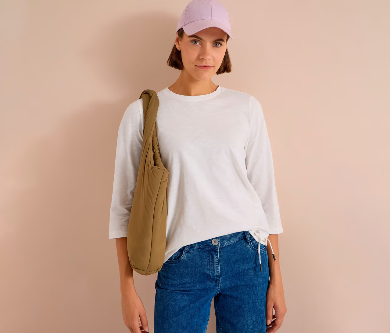 Femme portant une casquette violet clair, un T-shirt 3/4 CECIL blanc, un jean bleu et un sac à bandoulière matelassé kaki.