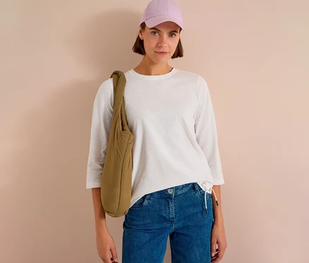 Femme portant une casquette violet clair, un T-shirt 3/4 CECIL blanc, un jean bleu et un sac à bandoulière matelassé kaki.