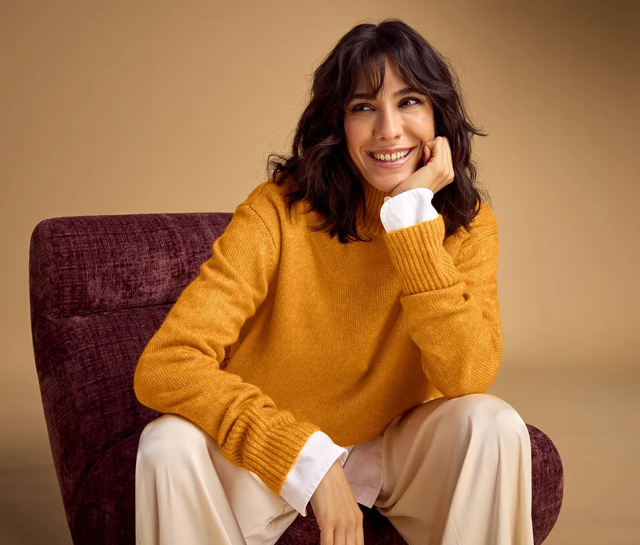 Une femme souriante aux cheveux foncés et ondulés est assise dans un fauteuil violet foncé, le menton posé sur la main. Elle porte un pull jaune sur une chemise blanche et un pantalon clair.