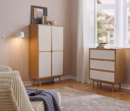 Salon lumineux avec une armoire Cabinetschrank à 2 portes et une commode à 3 tiroirs en bois et façades claires. Sur la commode se trouve une lampe de table à LED rechargeable Eglo « Mannera ». À gauche un canapé, à droite une fenêtre avec des rideaux.