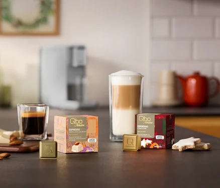 Une machine à café Qbo ICONIC Light Gold en arrière-plan. Sur un comptoir de cuisine se trouvent deux paquets de capsules Qbo Espresso, White Choc & Almond et Zimtstern, avec des capsules, un espresso et un latte macchiato, des amandes, du chocolat blanc et des biscuits Zimtstern.