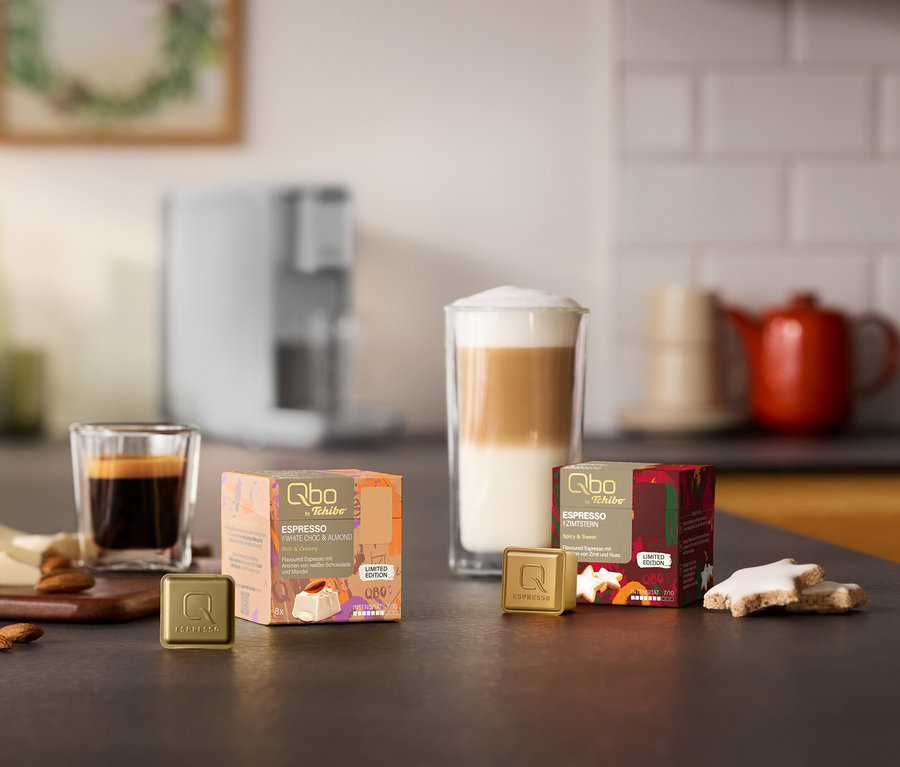 Une machine à café Qbo ICONIC Light Gold en arrière-plan. Sur un comptoir de cuisine se trouvent deux paquets de capsules Qbo Espresso, White Choc & Almond et Zimtstern, avec des capsules, un espresso et un latte macchiato, des amandes, du chocolat blanc et des biscuits Zimtstern.