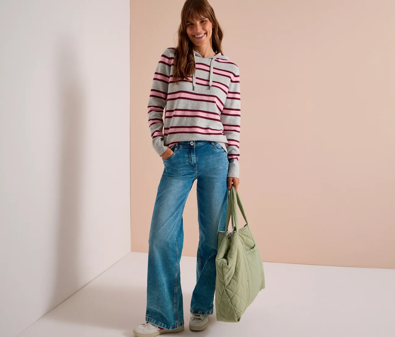 Une femme souriante se tient les mains dans les poches, vêtue d'un sweatshirt à capuche en maille CECIL gris à rayures roses et rouge foncé, d'un jean large bleu et de baskets blanches, tenant un sac fourre-tout matelassé vert clair.