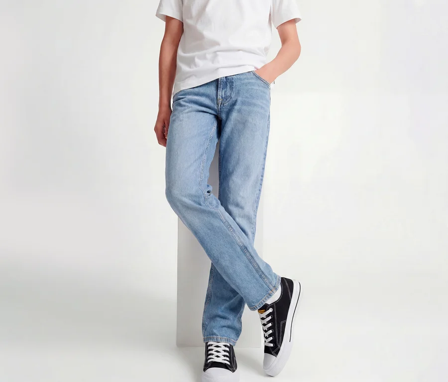 Personne portant un t-shirt blanc, un jean Jack&Jones JJOriginal Chris bleu clair et des baskets noires, appuyée contre un bloc blanc, jambes croisées.