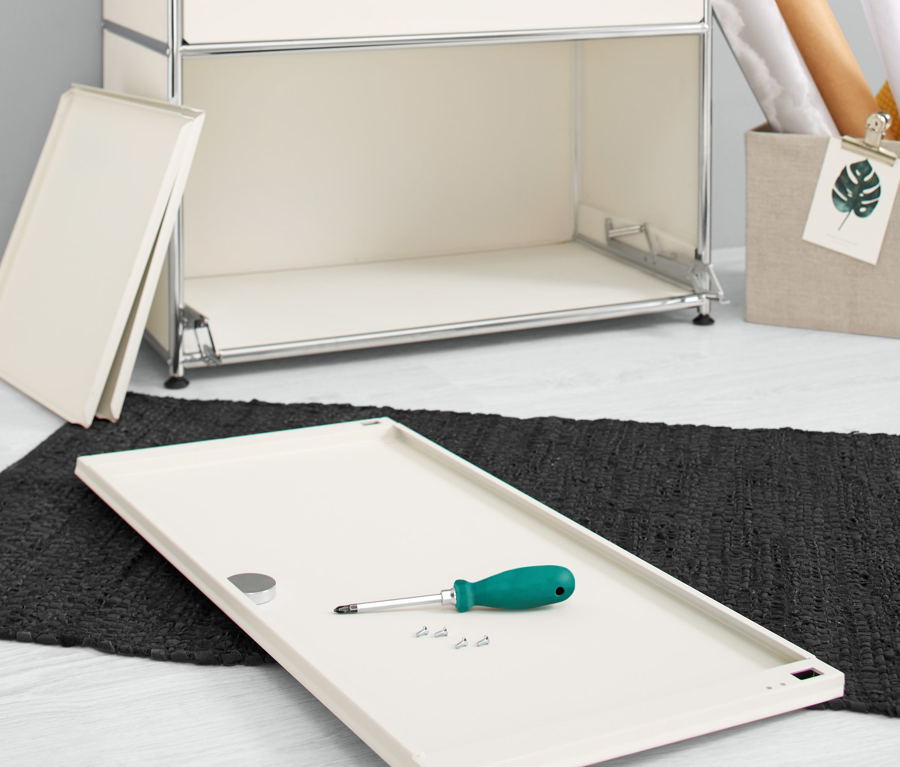 Un jeu d'abattants supplémentaires en métal « CN3 », blanc, repose sur le sol avec un tournevis et des vis à côté d'une commode.