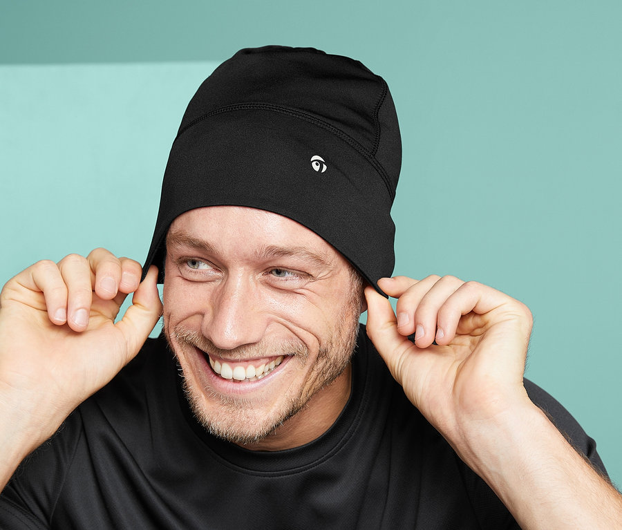 Un homme souriant tient les bords de son bonnet fonctionnel unisexe noir.