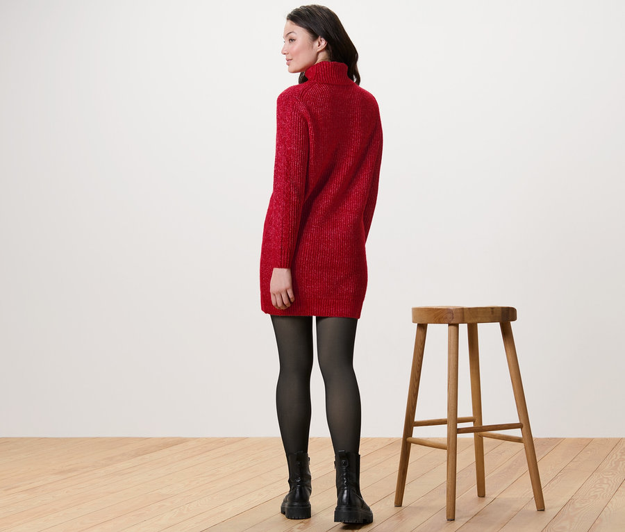 Femme portant un pull en maille longue avec torsades rouge, des collants noirs et des bottes, debout à côté d'un tabouret en bois.