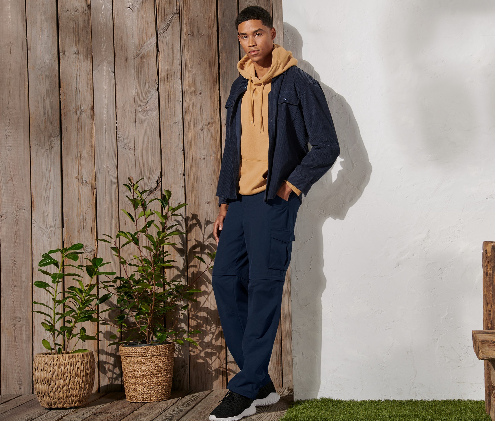 Un homme debout porte un pantalon fonctionnel 2 en 1 bleu marine, un sweat à capuche jaune et une veste.