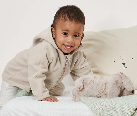 Un bébé souriant rampe sur un lit blanc et porte une veste en polaire pour bébé.
