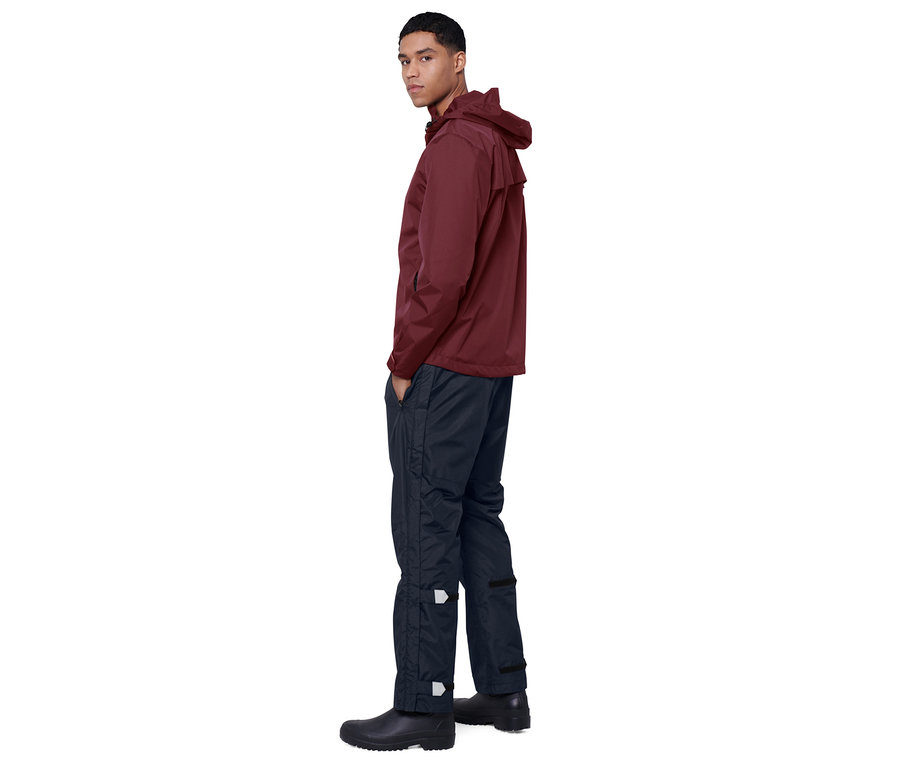 Un homme pose en veste bordeaux et pantalon de pluie unisexe high tech bleu marine.