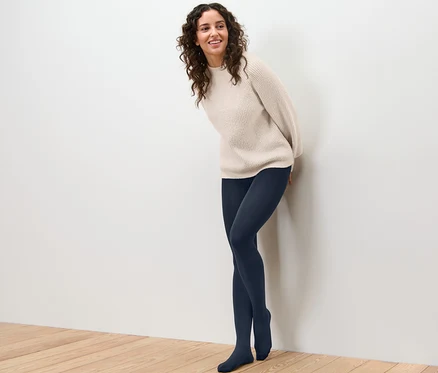 Une femme souriante appuyée contre un mur porte un collant thermique et un pull.