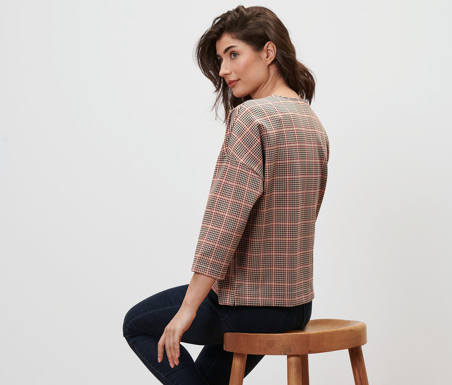 Femme assise sur un tabouret en bois, portant un sweat-shirt manches 3/4, pied de poule.