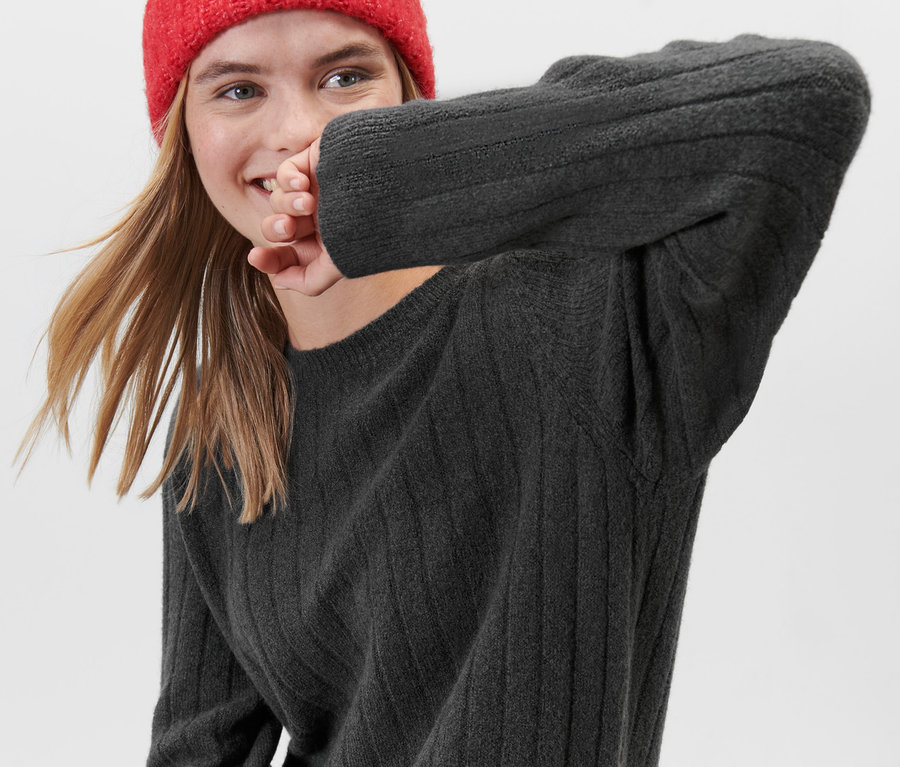 Une femme souriante portant une robe en tricot côtelée grise et un bonnet rouge.