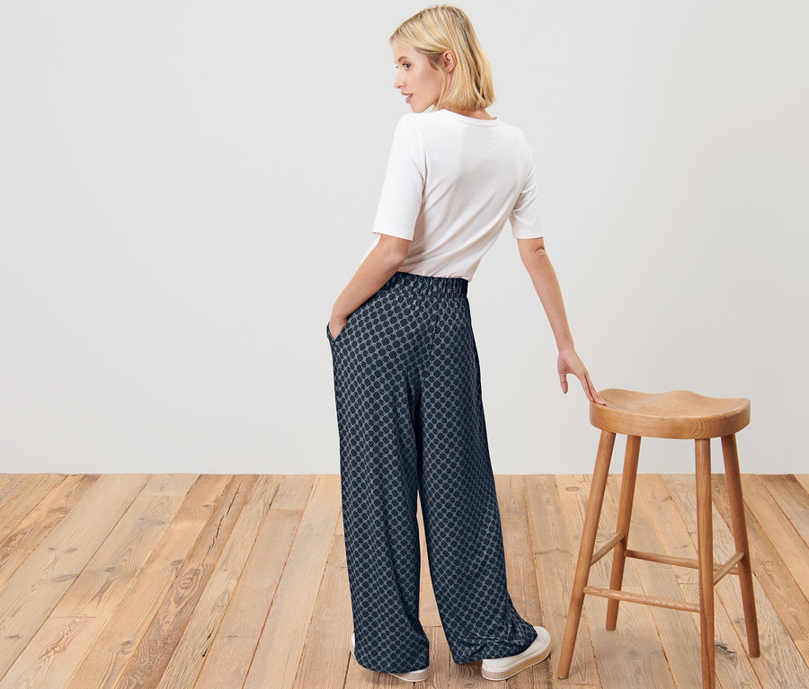 Femme blonde posant en pantalon palazzo imprimé bleu foncé et t-shirt blanc à côté d'un tabouret en bois.