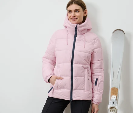 Une femme se tient à côté de skis et porte une veste de ski matelassée rose.