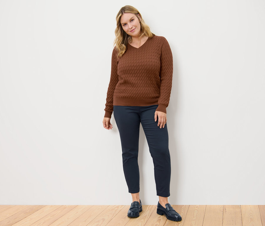 Une femme pose, portant un pull en maille torsadée cognac et un pantalon stretch bleu foncé.