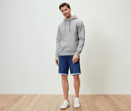 Un homme debout portant un sweat à capuche gris chiné, un short en jean et des baskets blanches.