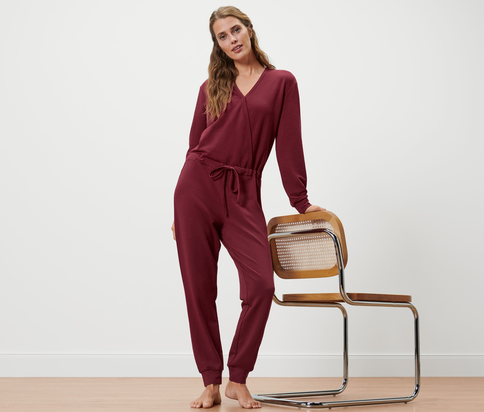Femme posant en combinaison d’intérieur bordeaux, une main sur une chaise.