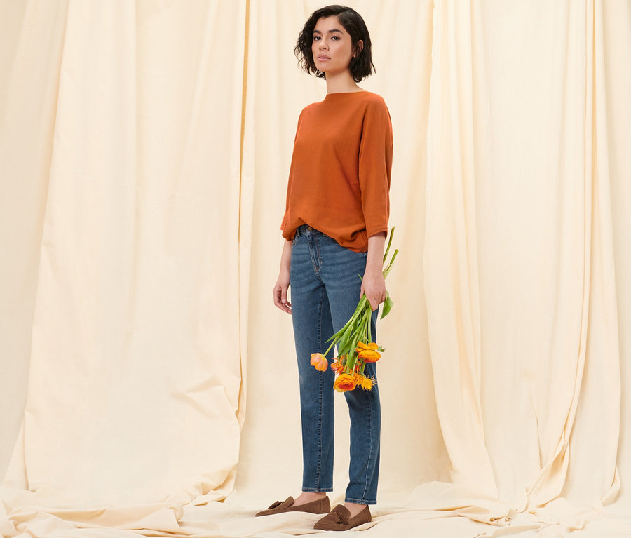 Une femme pose devant un fond en tissu en tenant des tulipes. Elle porte un pull en maille fine orange, un jean slim en denim « Fit Emma » bleu moyen et des mocassins en cuir velours.