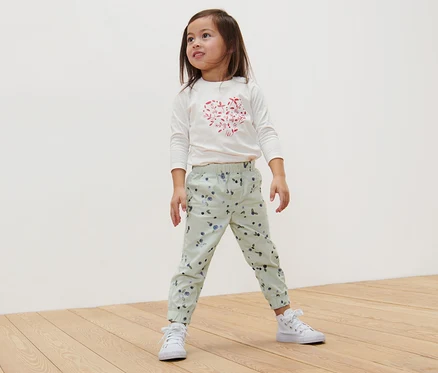 Une fille se tient sur un plancher en bois et porte un pantalon à enfiler pour enfant – Coupe «	Ella	», myrtilles.