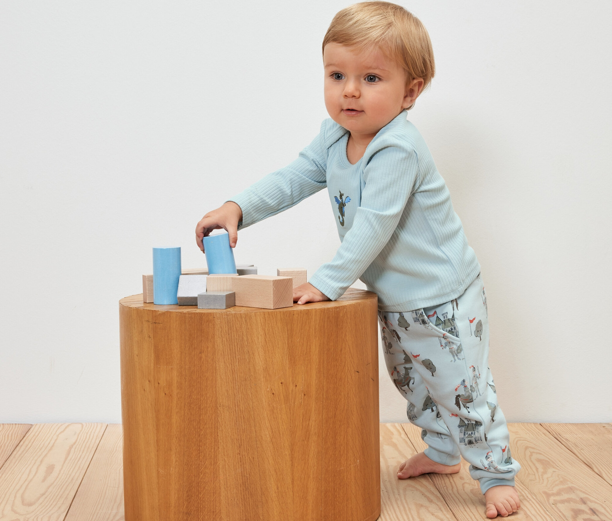 Un bébé joue avec des blocs de bois sur une table en bois, portant une tenue sur le thème des chevaliers et 2 pantalons de survêtement pour bébé, chevalier.