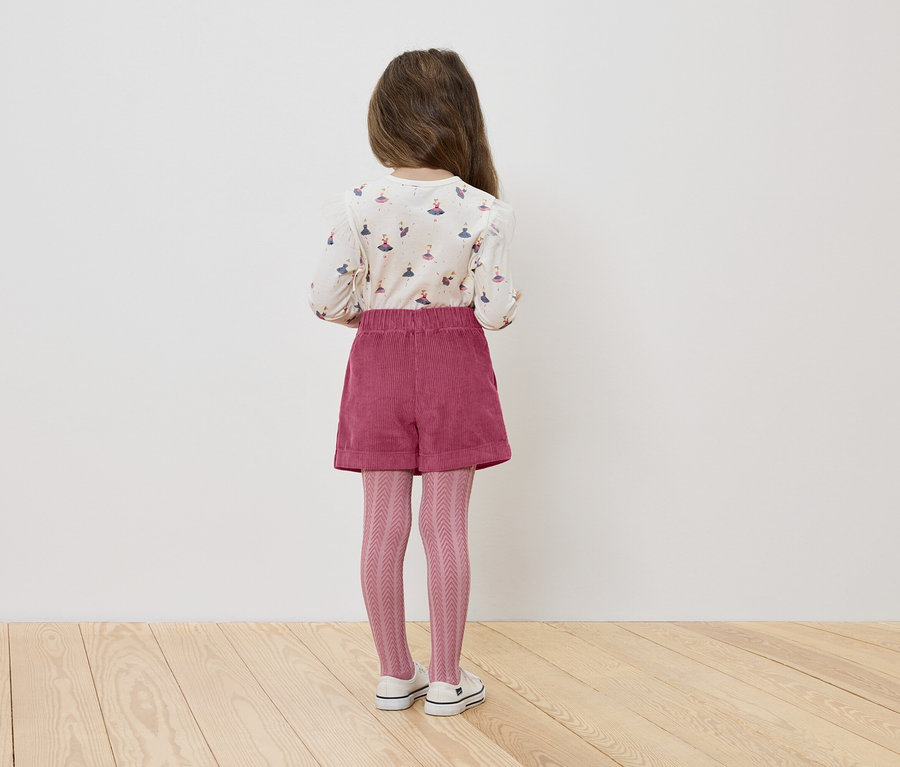 Une fillette, vue de dos, porte un t-shirt avec des ballerines, un short rose et des collants tricotés.