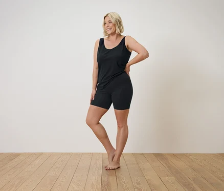 Une femme blonde en débardeur noir et short pose sur un plancher en bois.
