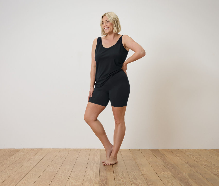 Une femme blonde en débardeur noir et short pose sur un plancher en bois.