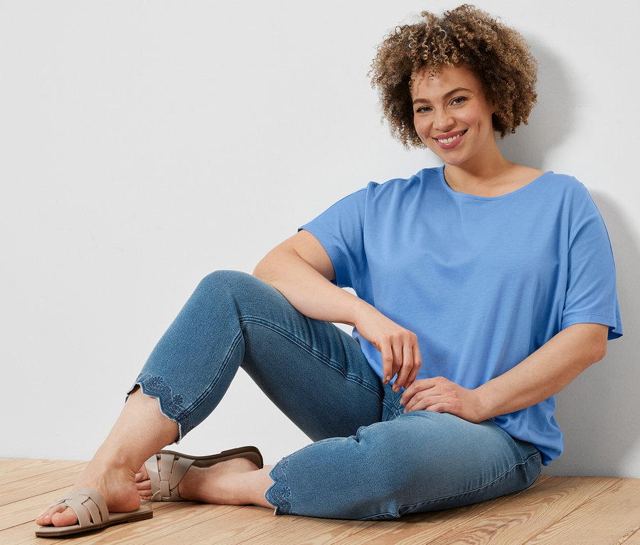 Femme assise sur un plancher en bois portant une blouse bleu clair et un tregging 7/8 à broderie ajourée.
