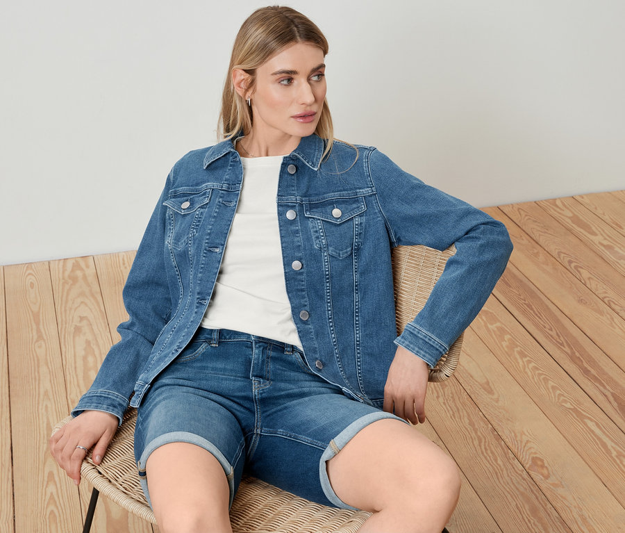 Une femme est assise sur une chaise et porte une veste en jean, un t-shirt basique blanc et un bermuda en jean.