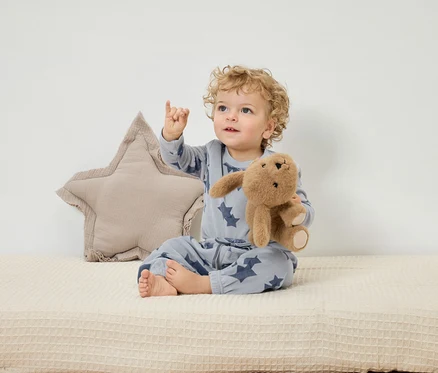 Un enfant est assis sur un tapis beige, tenant un lapin en peluche et pointant son doigt vers le haut. Derrière lui se trouve un coussin en forme d'étoile.