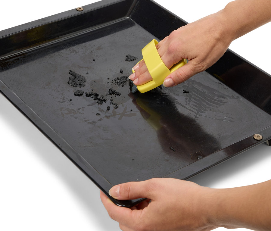 Des mains nettoient une plaque de cuisson noire avec un outil de nettoyage jaune.
