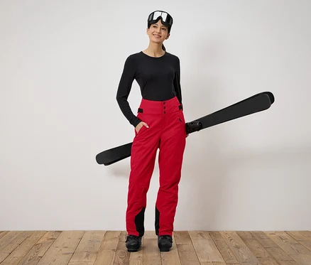 Une femme debout tenant des skis. Elle porte un haut noir à manches longues, un pantalon rouge et des lunettes de ski.