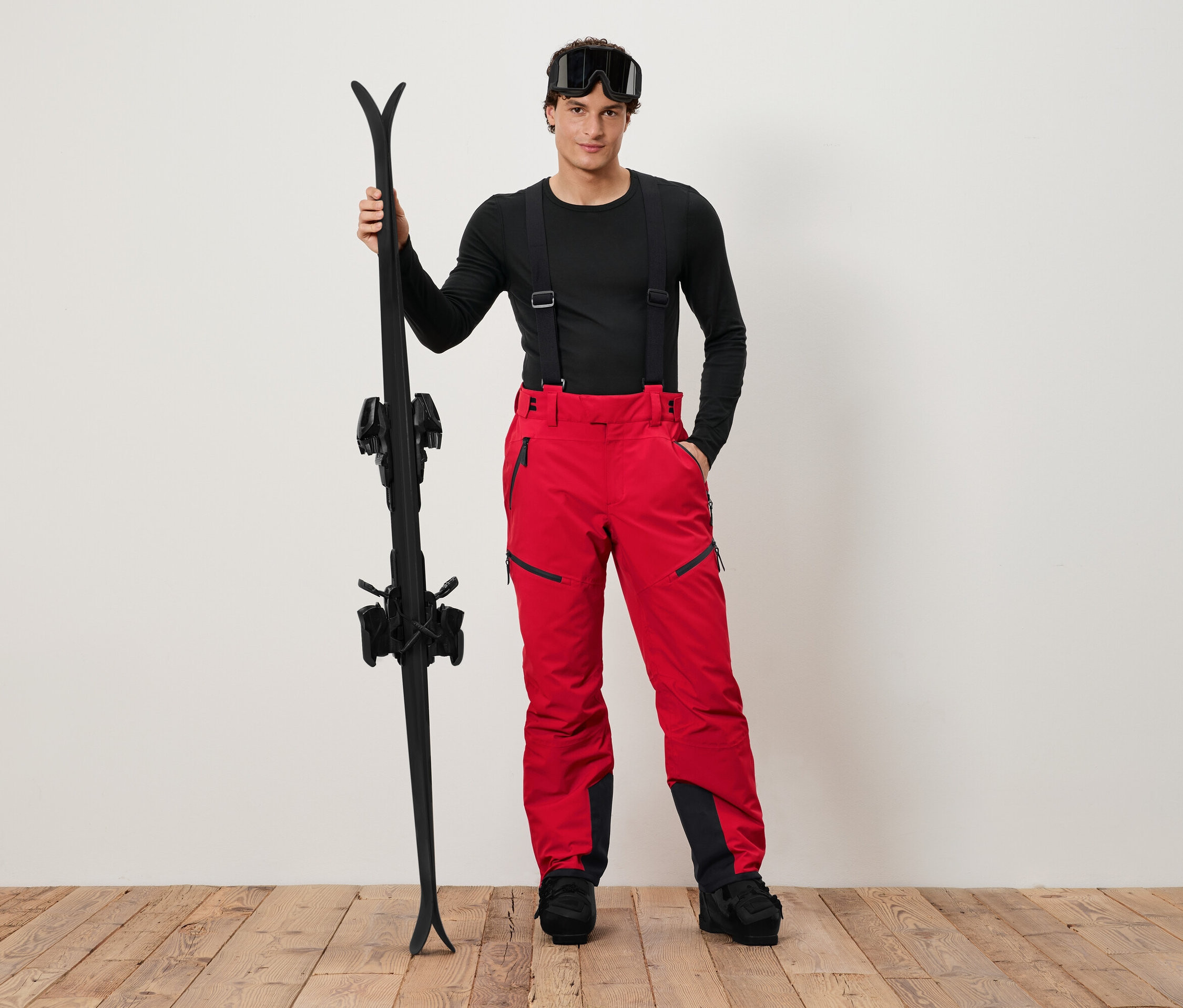 Un homme en chemise noire et pantalon de ski rouge tenant des skis noirs.