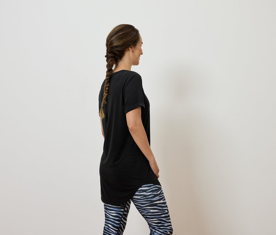 Une femme avec des cheveux bruns tressés en une natte se tient dos à l'appareil photo et porte un t-shirt noir et un legging bleu et blanc.