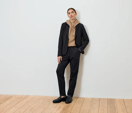 Une femme pose portant un blazer noir, un sweat à capuche marron et un pantalon noir.
