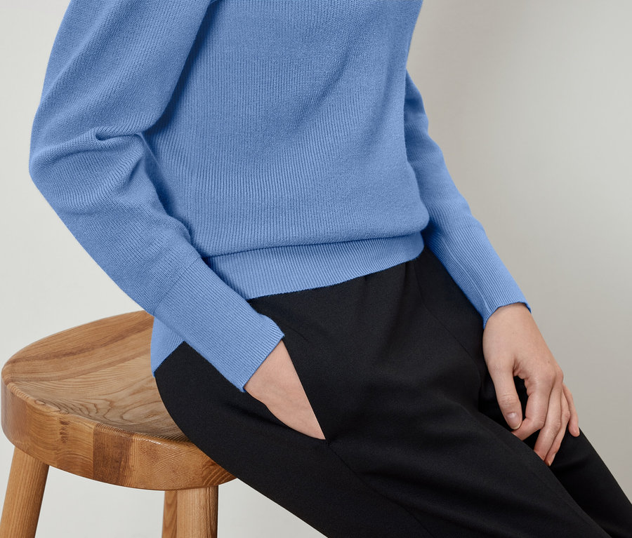 Femme assise sur un tabouret en bois, portant un pull bleu et un pantalon noir avec la main dans la poche.