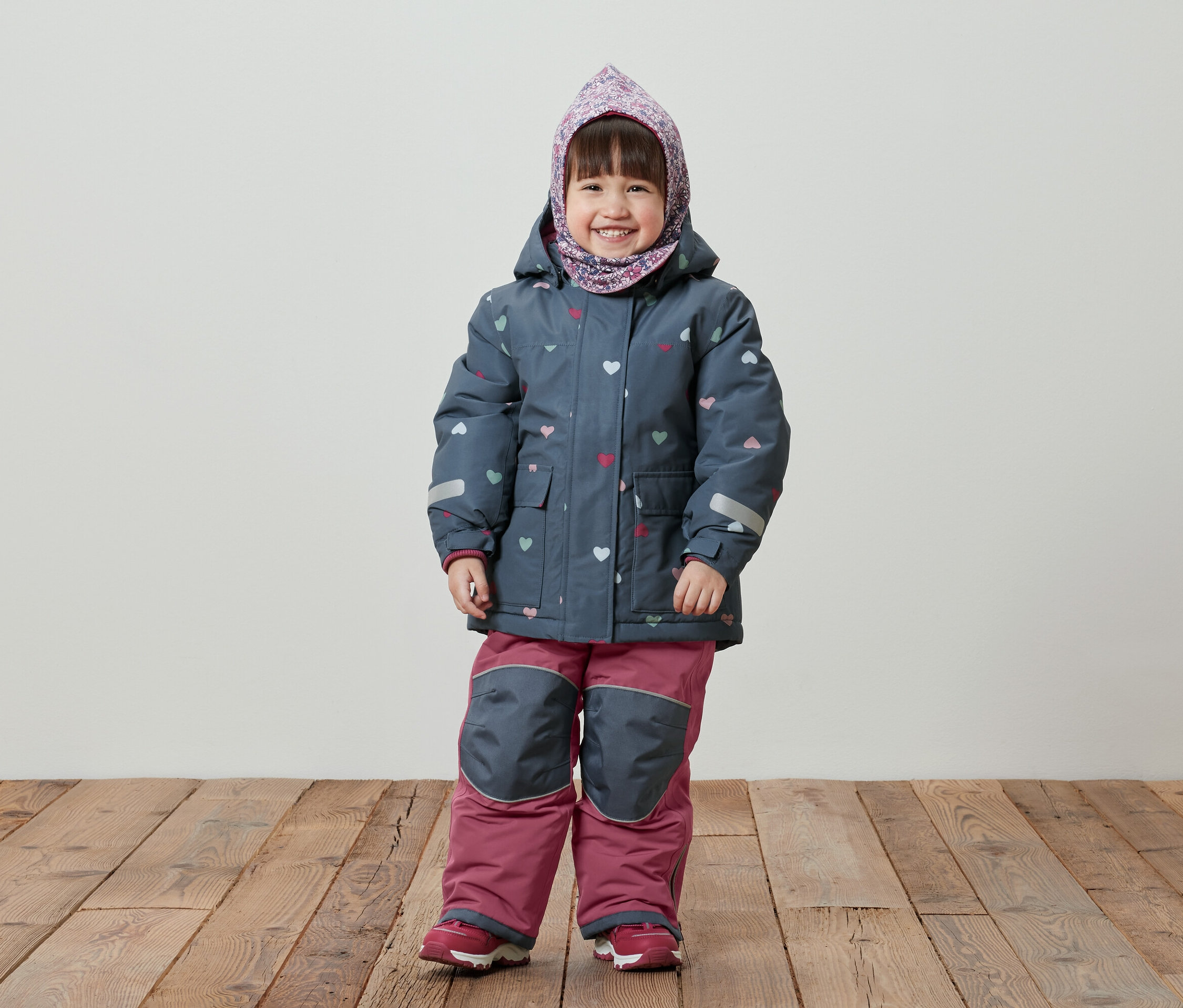 Une petite fille souriante se tient sur un plancher en bois, portant une veste d'hiver bleue à motifs de cœurs, un pantalon rose et un foulard sur la tête.