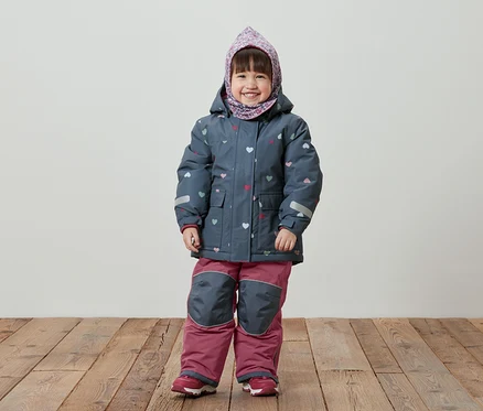 Une petite fille souriante se tient sur un plancher en bois, portant une veste d'hiver bleue à motifs de cœurs, un pantalon rose et un foulard sur la tête.
