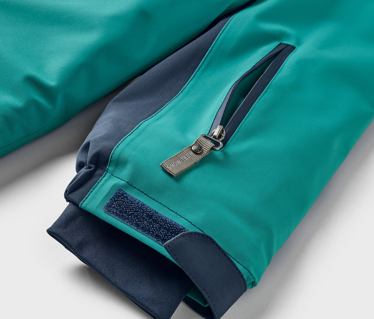 Gros plan sur une veste turquoise avec des détails bleu foncé, montrant une poche zippée et une fermeture velcro sur la manche.